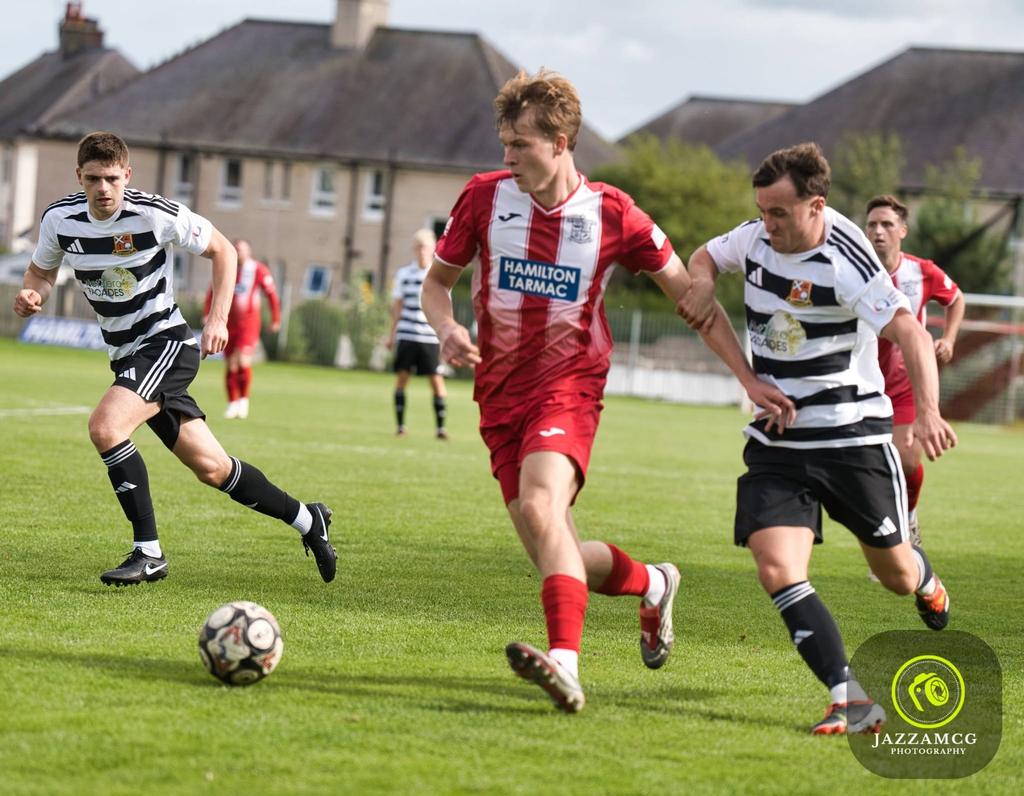 Hurlford United midfielder in the red Hamilton Tarmac shirt chasing the ball between two opponents